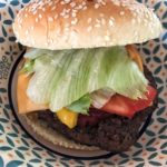 Aufnahme eines Cheeseburgers mit Sesambrötchen, Salat, Tomate, geschmolzenem Käse und gegrilltem Patty auf einem gemusterten Pappteller beim Deutsch-Amerikanisches Volksfest Grafenwöhr. — Bild: volksfest-grafenwoehr.de