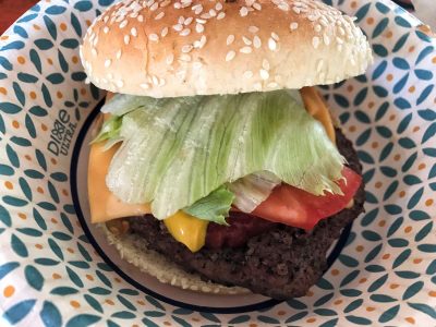 Aufnahme eines Cheeseburgers mit Sesambrötchen, Salat, Tomate, geschmolzenem Käse und gegrilltem Patty auf einem gemusterten Pappteller beim Deutsch-Amerikanisches Volksfest Grafenwöhr. — Bild: volksfest-grafenwoehr.de
